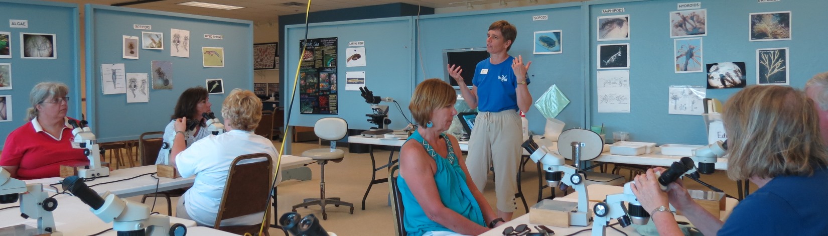 A teacher talking to adults who are seated at microscopes